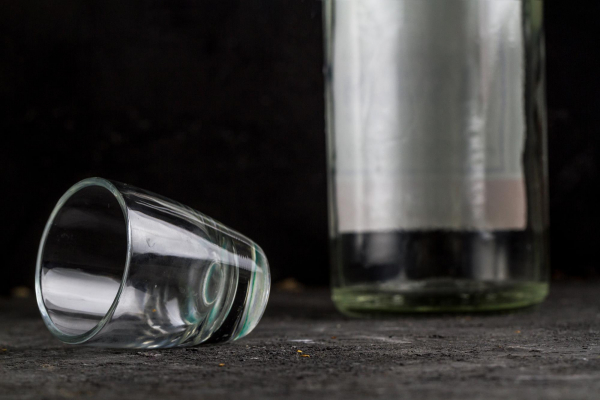 Na stole stoi pusta butelka po alkoholu, a obok leży przewrócony pusty kieliszek po wódce