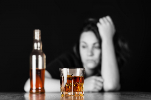 Przygnębiona kobieta z problemem alkoholowym siedzi przy stole. Przed nią stoi butelka i szklanka z alkoholem